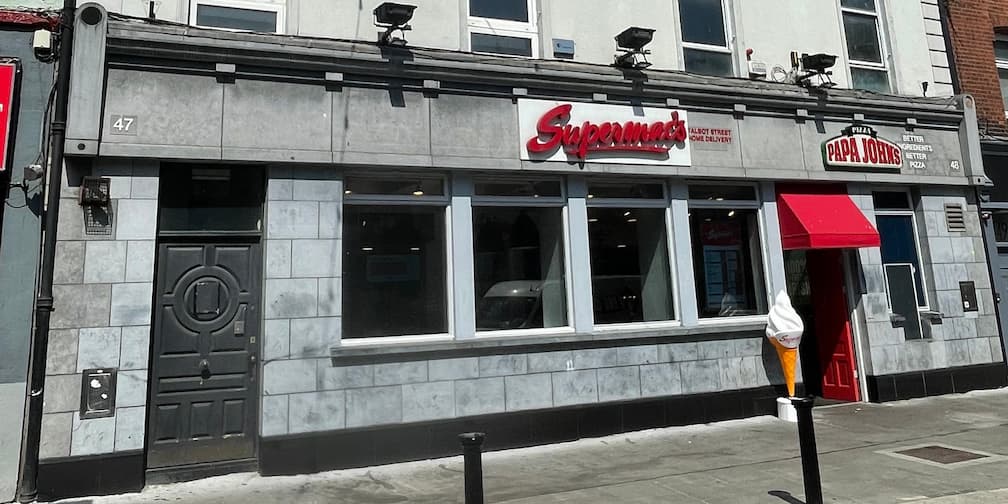 Supermac's and Papa John's restaurant exterior on Talbot Street, Dublin, Ireland. Features a grey stone facade, prominent "Supermac's" and "Papa John's Pizza" signage, red awning over Papa John's entrance, and a white ice cream cone sculpture.